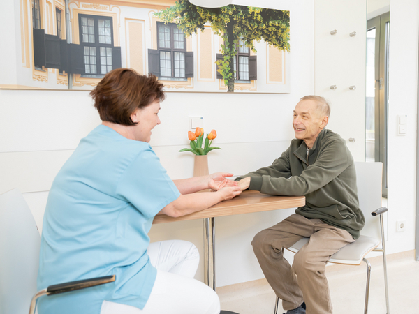 Pflegerin und Patient halten Hände an einem Tisch sitzend