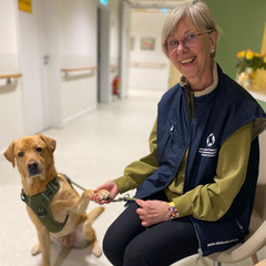 Eine ehrenamtlich engagierte Frau hält in einem Flur ihren Therapiehund an der Leine und blickt lächelnd in die Kamera im Diakonie Hospiz Woltersdorf.