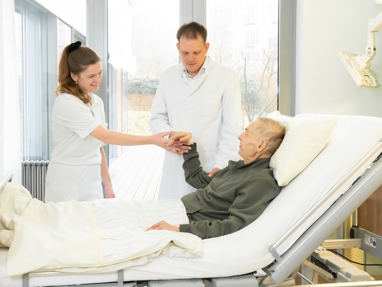 Ärztin und Arzt stehen an Bett mit einem Patienten und die Ärztin hält die Hand