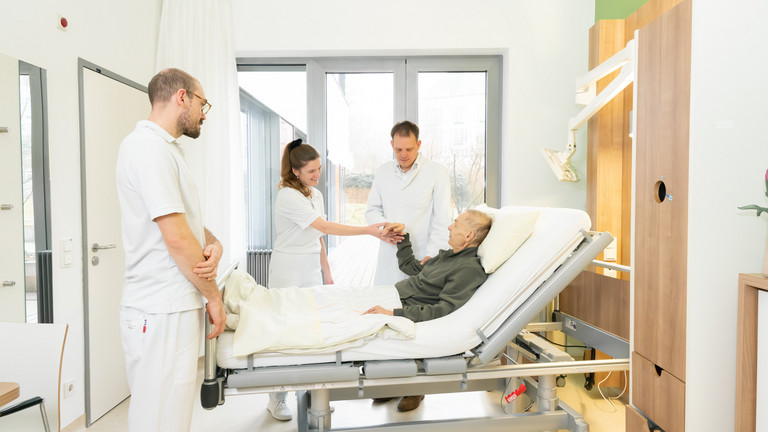 Ärztin und zwei Ärzte stehen an Bett mit einem Patient und die Ärztin hält die Hand