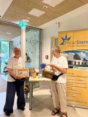 Zwei ehrenamtlich engagierte Frauen stehen mit Prospekten und einem Korb vor einem Informationsstand mit Informationsmaterialien und großem Banner des Vereins PolarStern Neuenhagen e.V. im Foyer des Diakonie Hospiz Woltersdorf.