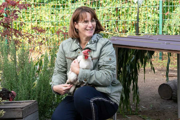 Eine ehrenamtlich engagierte Frau hält lachend ein weißes Huhn in den Armen im Hühnergarten mit Hühnervoliere auf dem Gelände des Diakonie Hospiz Woltersdorf.