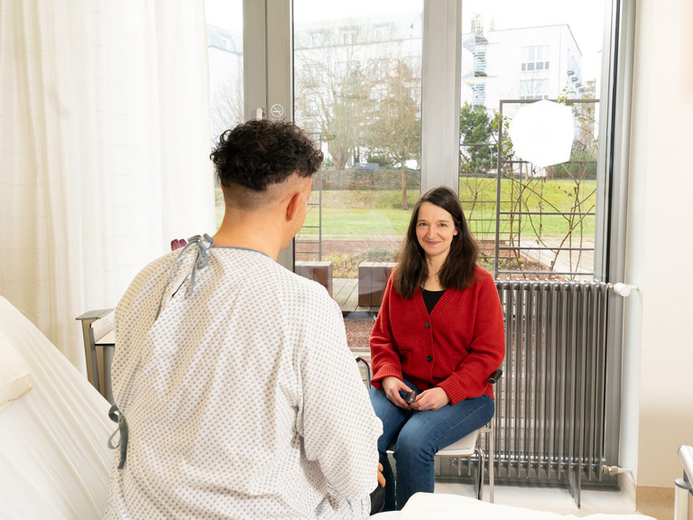 Mitarbeiterin im Gespräch mit Patient, der auf einem Bett sitzt
